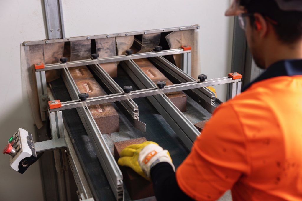 A worker in high-visibility orange and blue clothing and a white helmet operates a large industrial brick-cutting machine. He is standing in front of a Ferrari & Cigarini machine, performing quality control on the bricks.