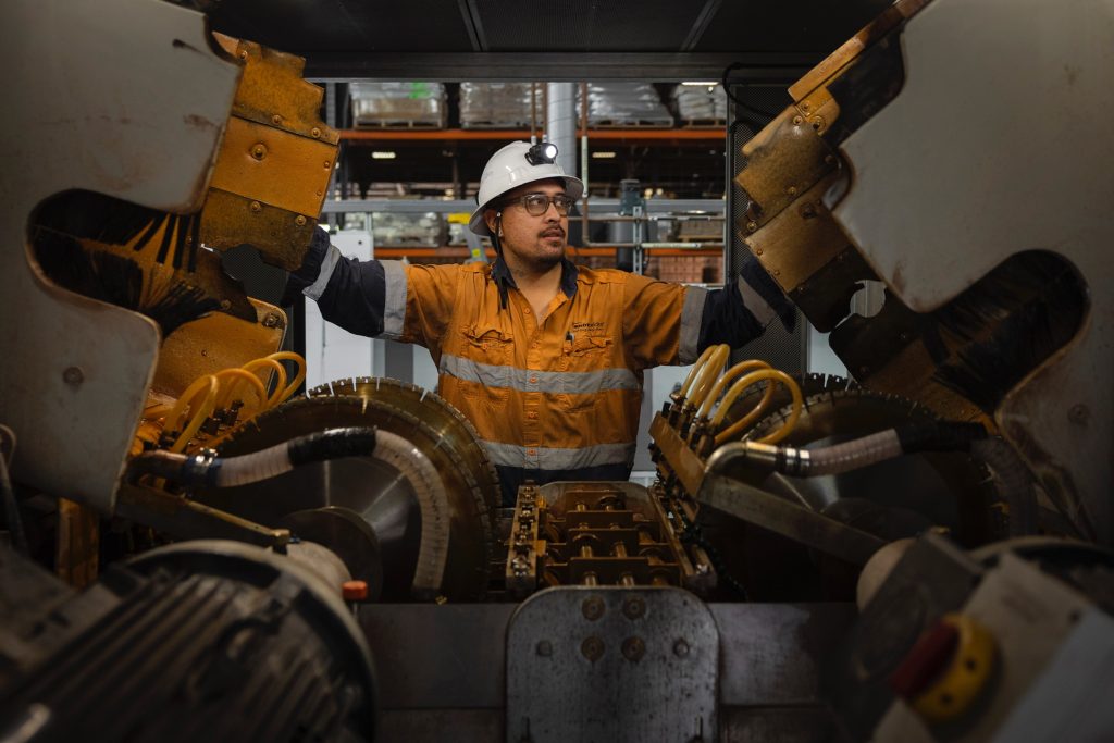 A worker in high-visibility orange and navy clothing and a white helmet operates a large industrial brick-cutting machine. He is standing between two open mechanical panels, with complex cutting blades and hydraulic tubes visible around him. The background shows shelves with materials inside a factory setting